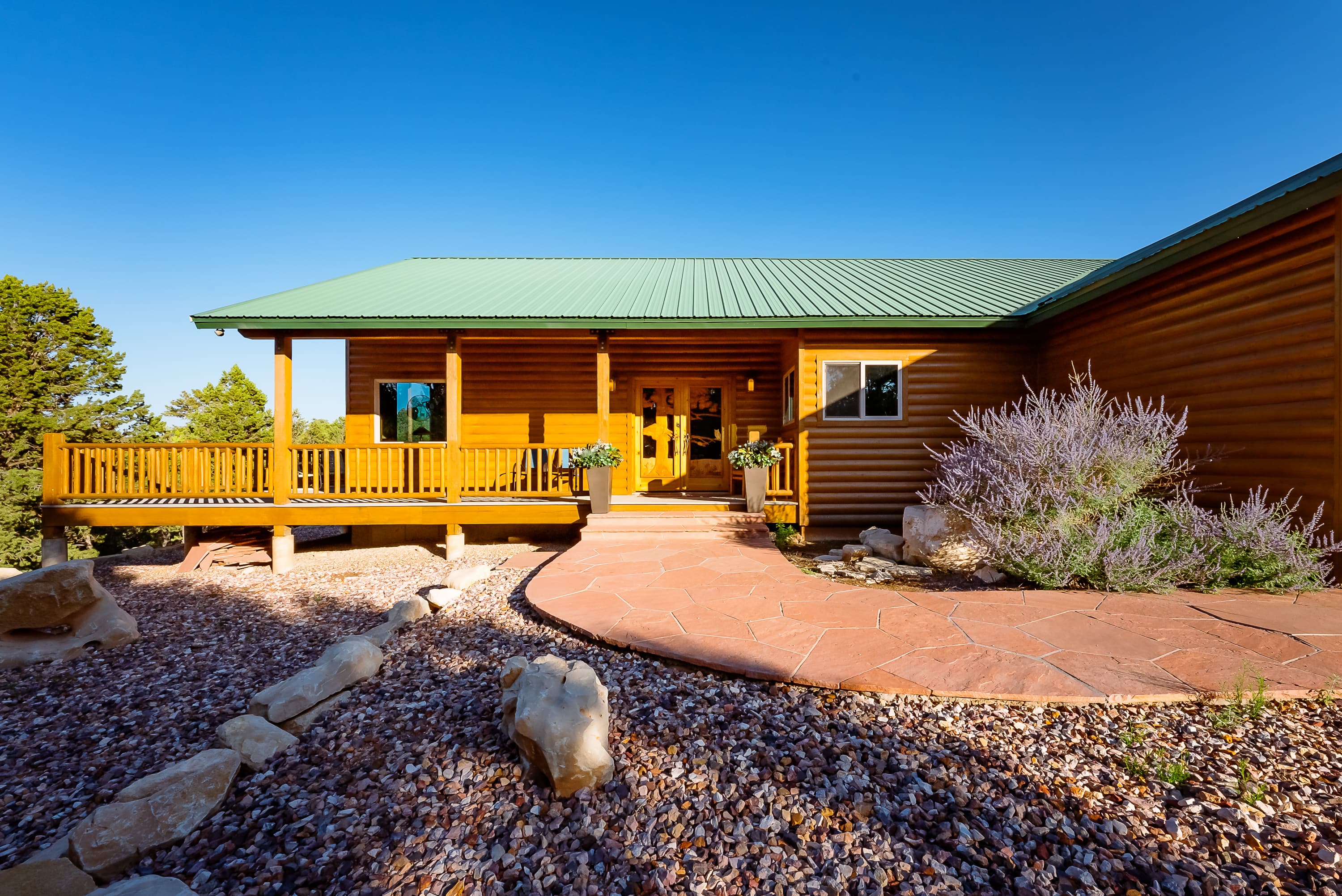 A wooden house with a green metal roof, featuring a front porch and landscaped stone pathway.