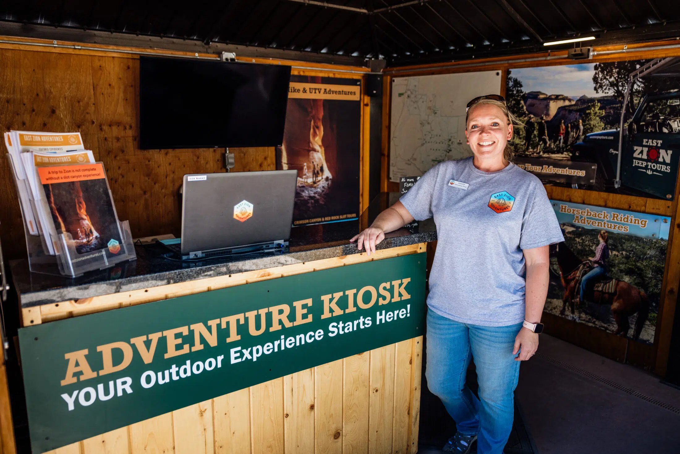 A woman stands behind an adventure kiosk with promotional materials and a computer, ready to assist customers.