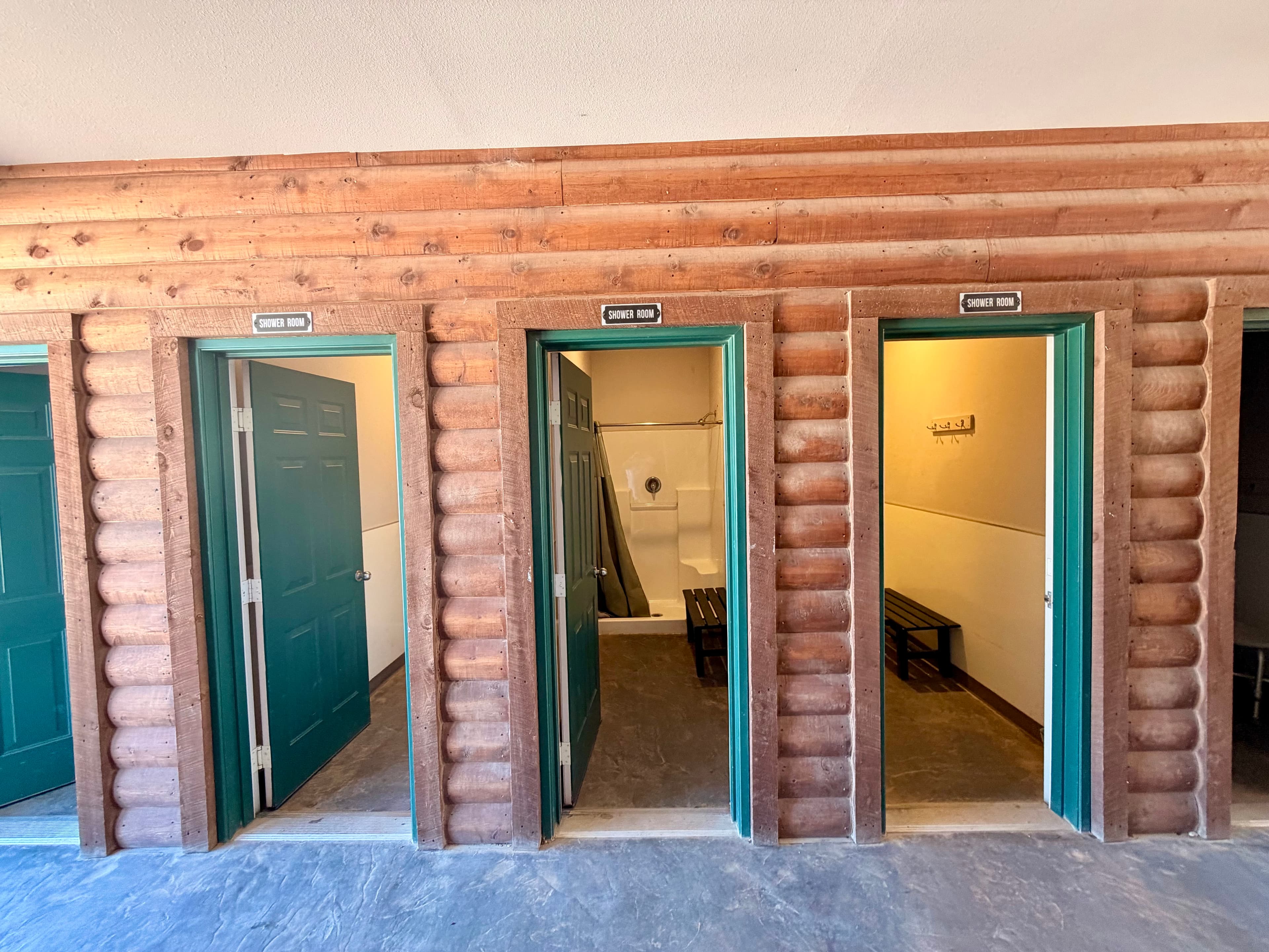 Three green doors lead to separate restroom or shower stalls against a wooden log wall.
