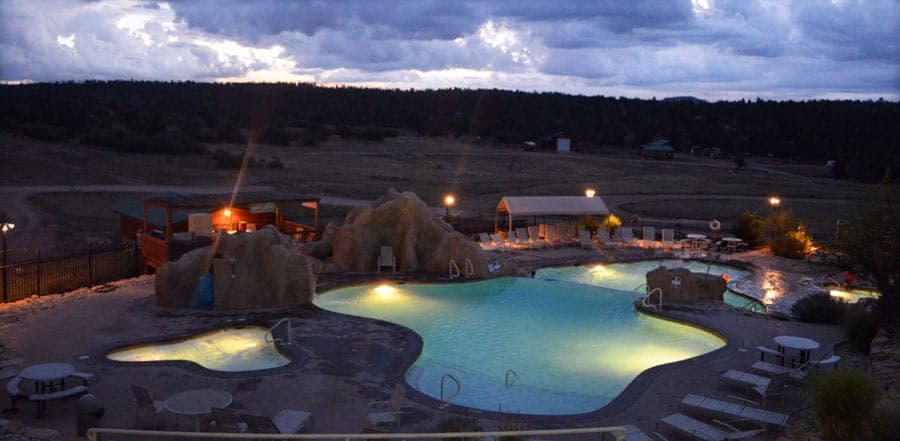 A nighttime view of a lit swimming pool surrounded by landscaped rocks and lounge chairs.