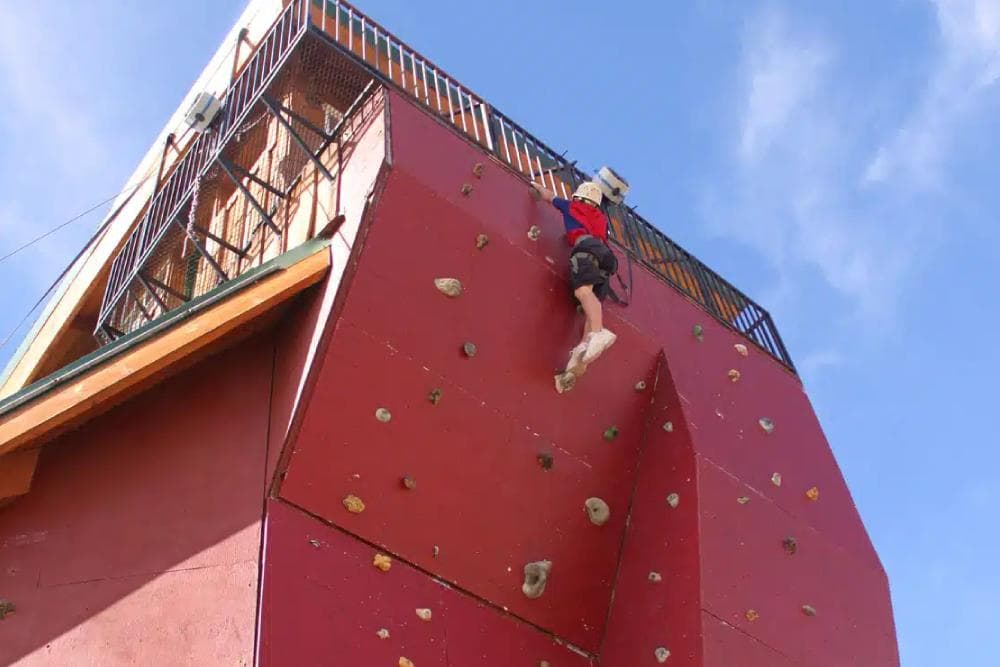 A climber ascends a red outdoor rock climbing wall under a clear blue sky.