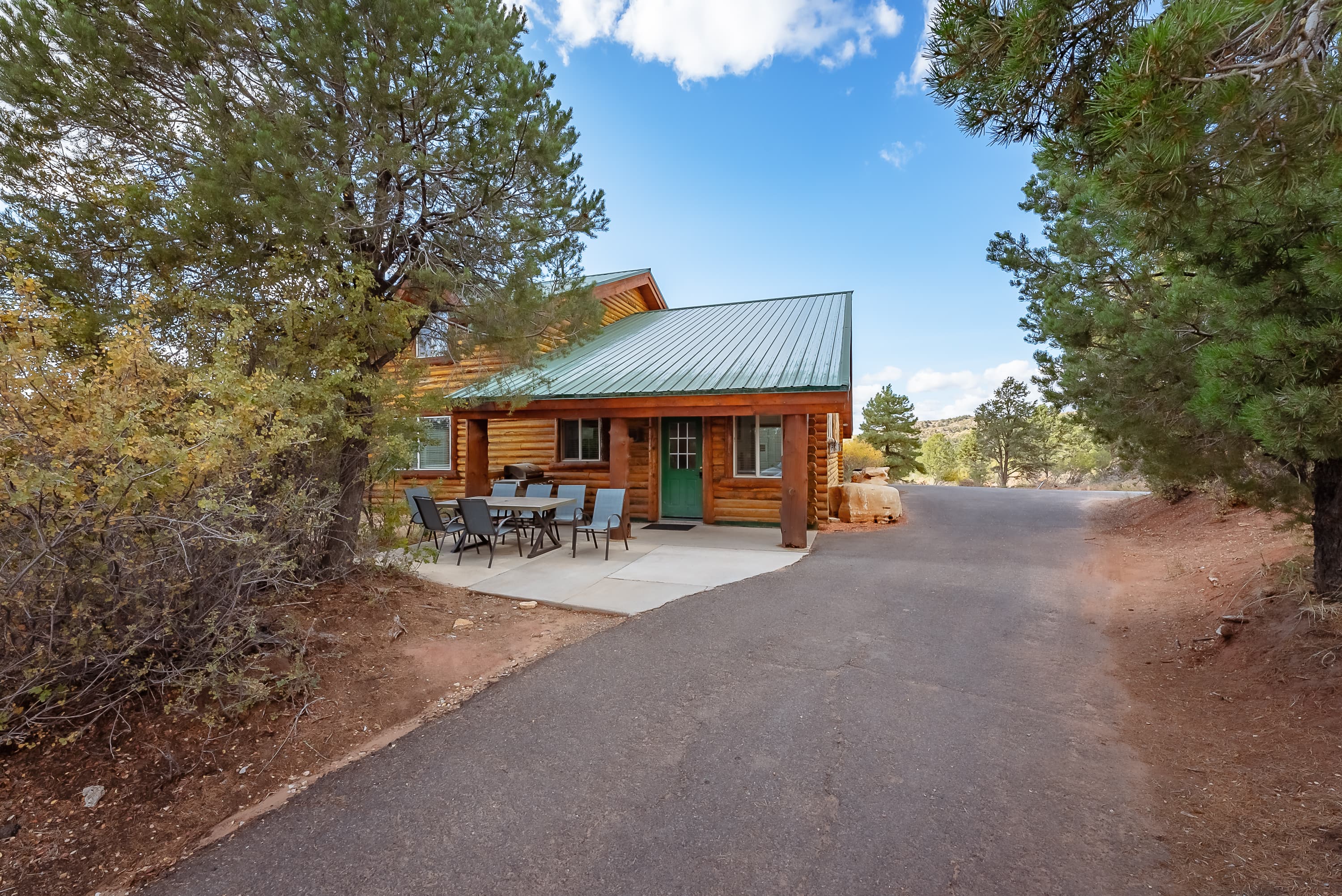 A rustic log cabin with a green metal roof surrounded by trees and a gravel driveway.