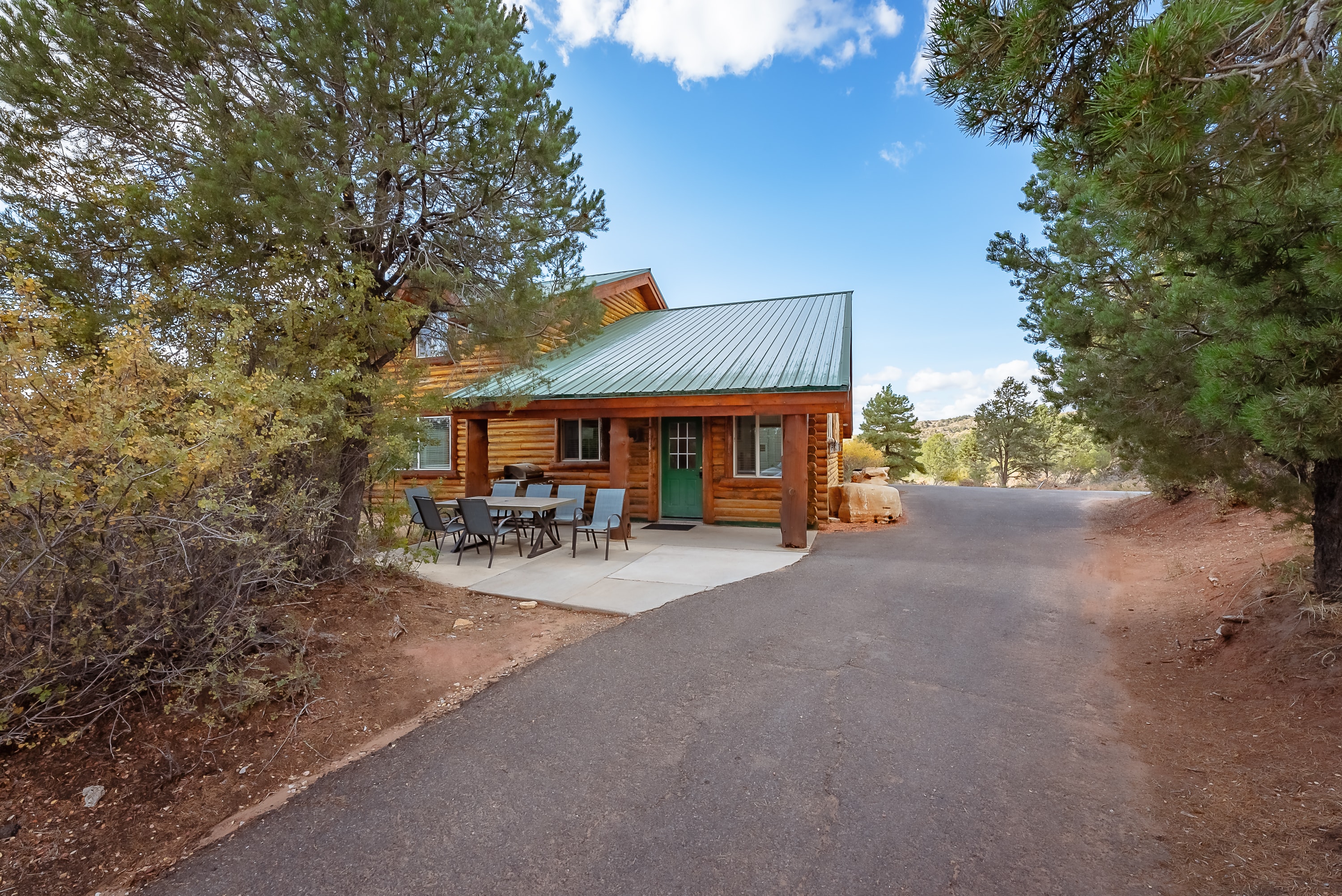 A rustic log cabin with a green metal roof surrounded by trees and a gravel driveway.