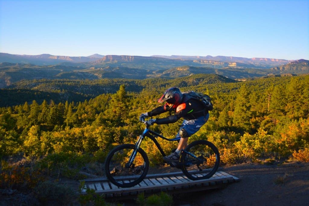 A mountain biker rides over a wooden bridge with a scenic backdrop of hills and forests.