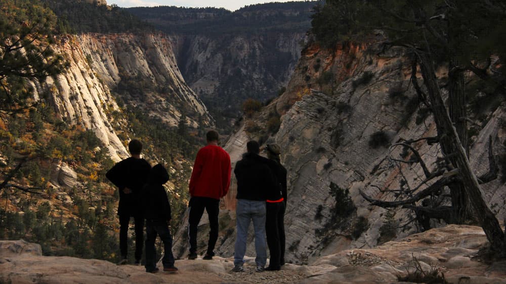 A group of four people stands on a rocky ledge, overlooking a deep canyon surrounded by rugged cliffs.