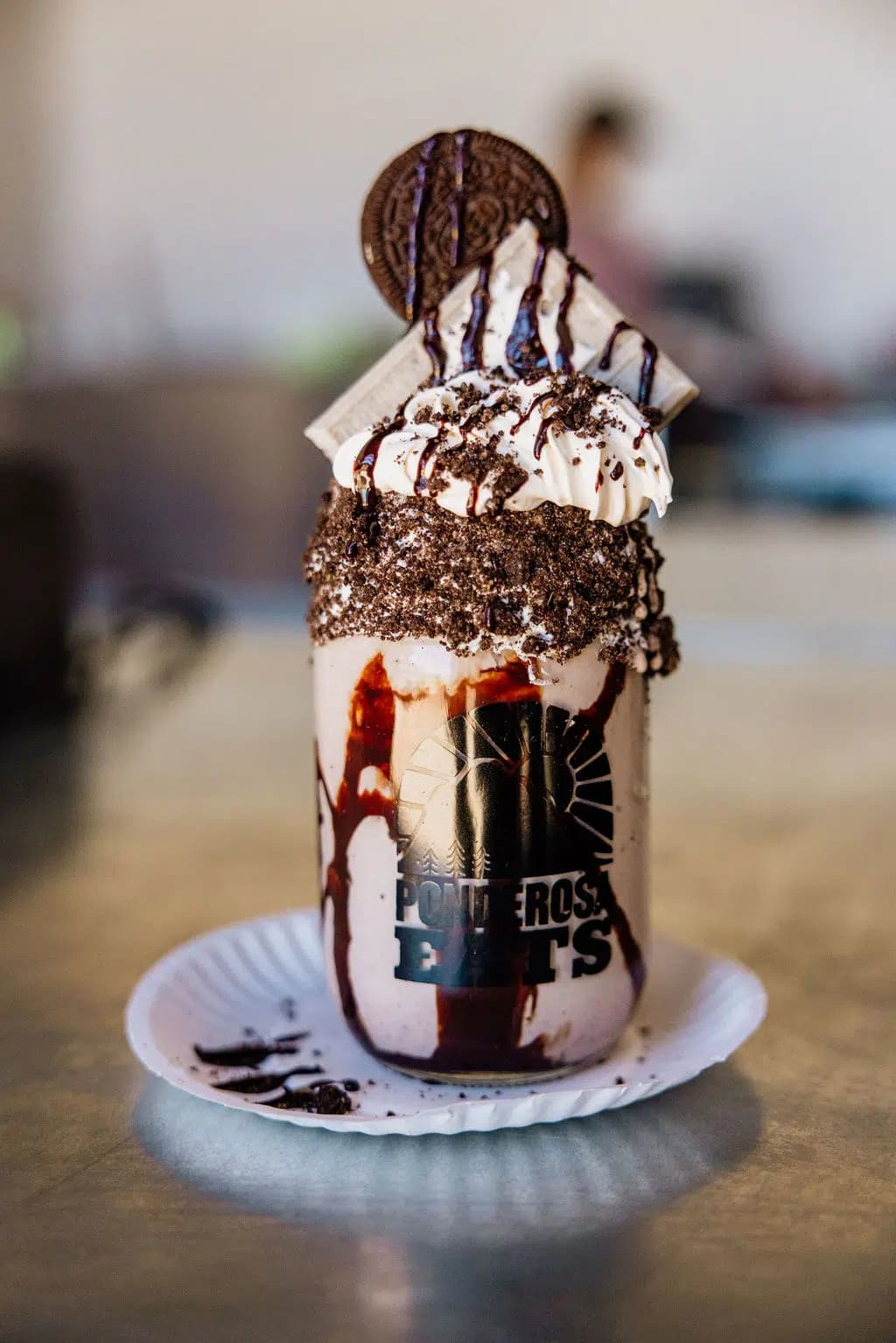 A decadent milkshake topped with whipped cream, chocolate drizzle, Oreos, and served in a branded glass.