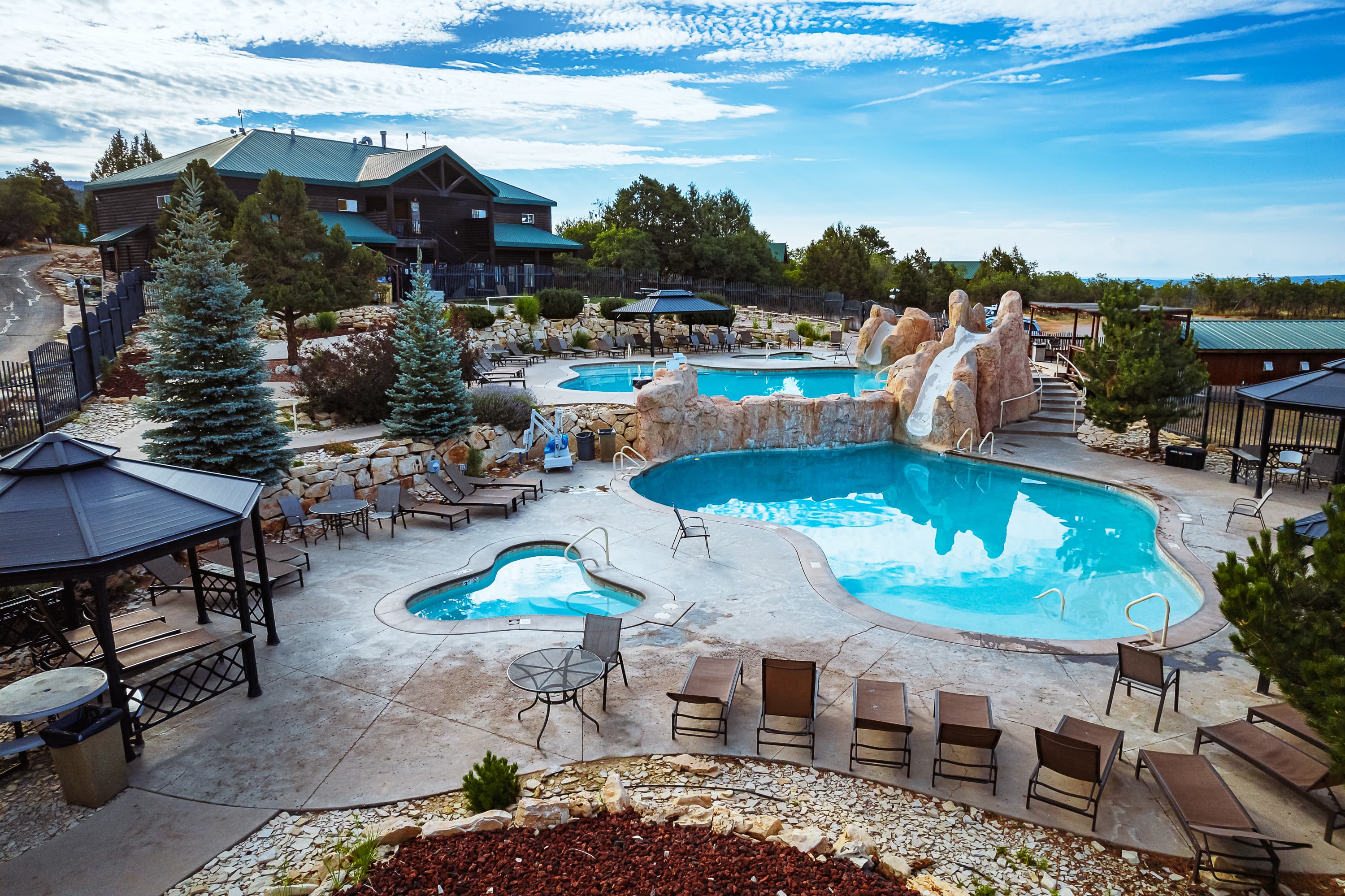 A scenic outdoor pool area featuring multiple swimming pools, lounge chairs, and a backdrop of trees and a blue sky.