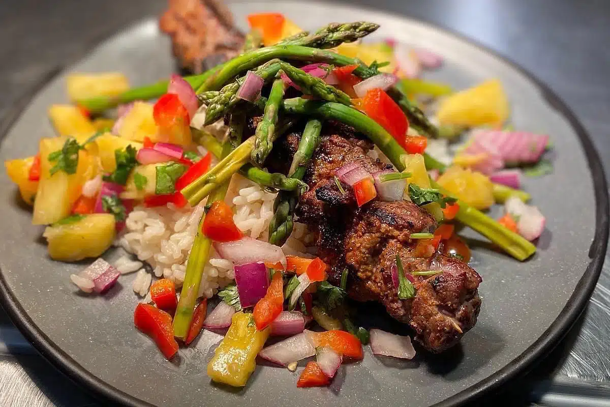 A plate of grilled meat skewers topped with asparagus and mixed vegetables, served over rice with pineapple.