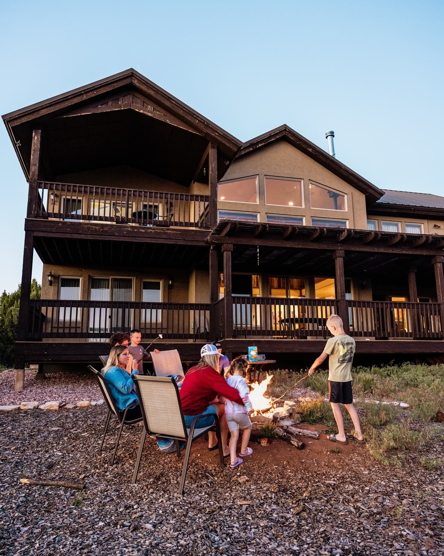 A group of people gathered around a fire pit in front of a large house at dusk.