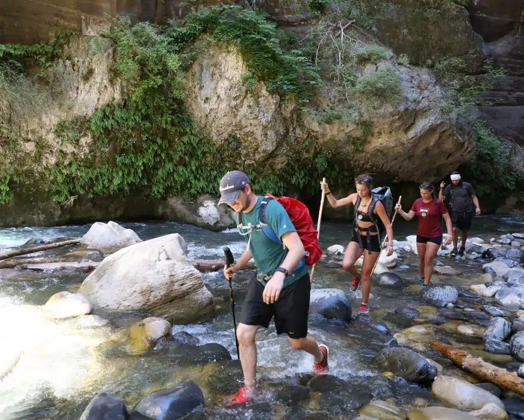 A group of hikers wades through a rocky riverbed surrounded by lush greenery.