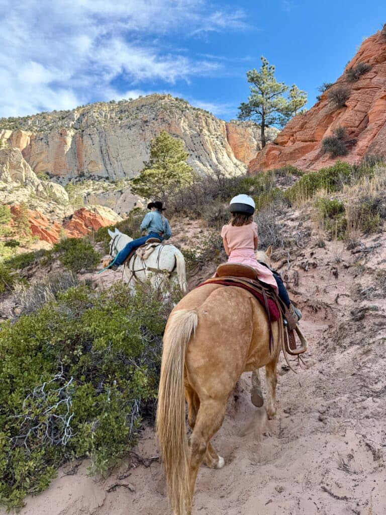 Two riders on horses navigate a rocky trail surrounded by colorful cliffs and greenery.