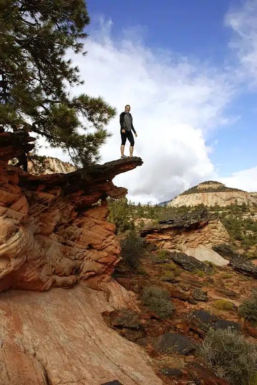 A hiker stands on a rocky ledge overlooking a scenic landscape with clouds in the sky.