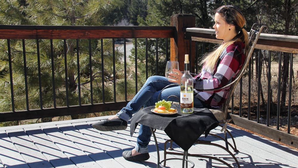 A woman sits on a porch chair, enjoying a drink and a snack with a scenic backdrop of trees.