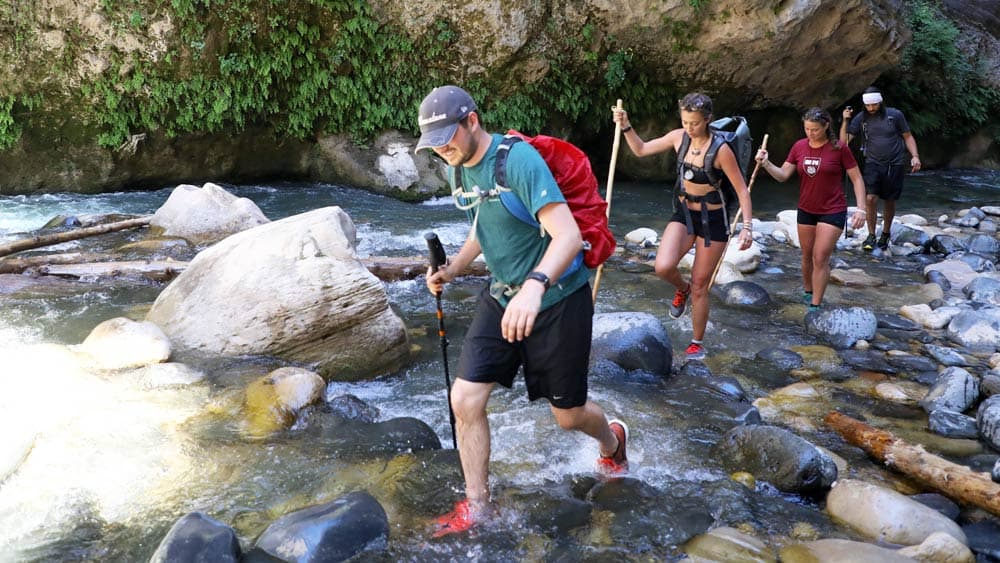 A group of hikers wades through a rocky stream surrounded by greenery.