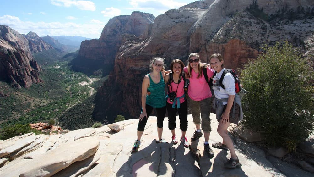 Four women pose together on a rocky ledge with a scenic canyon backdrop.