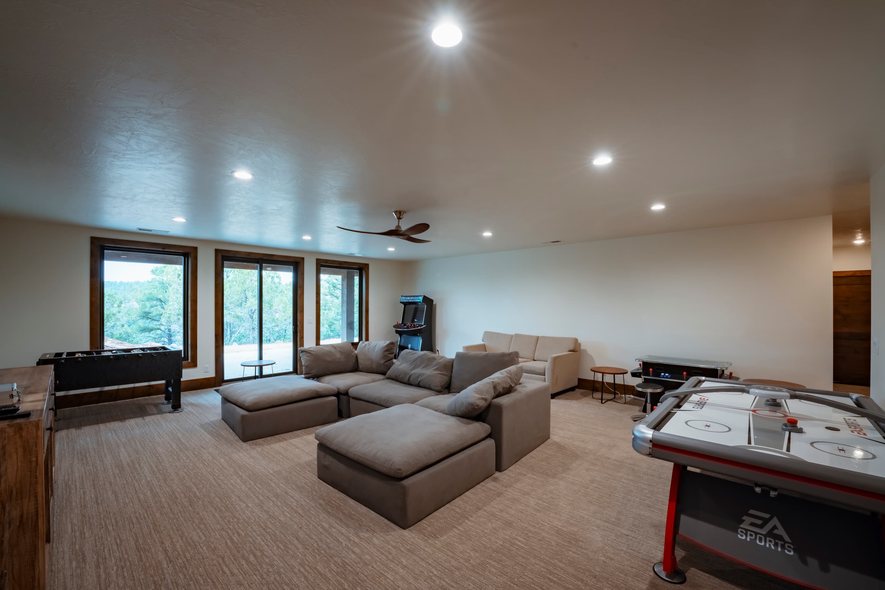 A spacious, well-lit living area featuring a large sectional sofa, air hockey table, and large windows.
