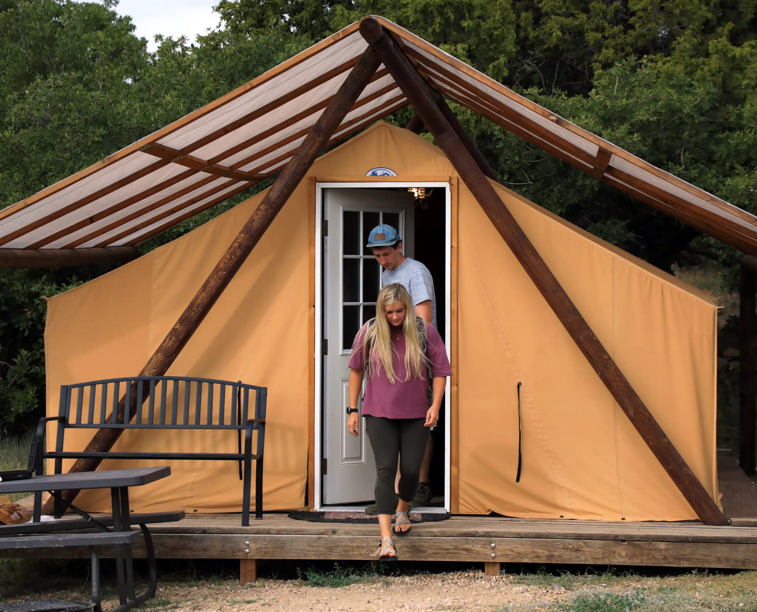 A man and woman step out of a tan glamping tent surrounded by greenery.