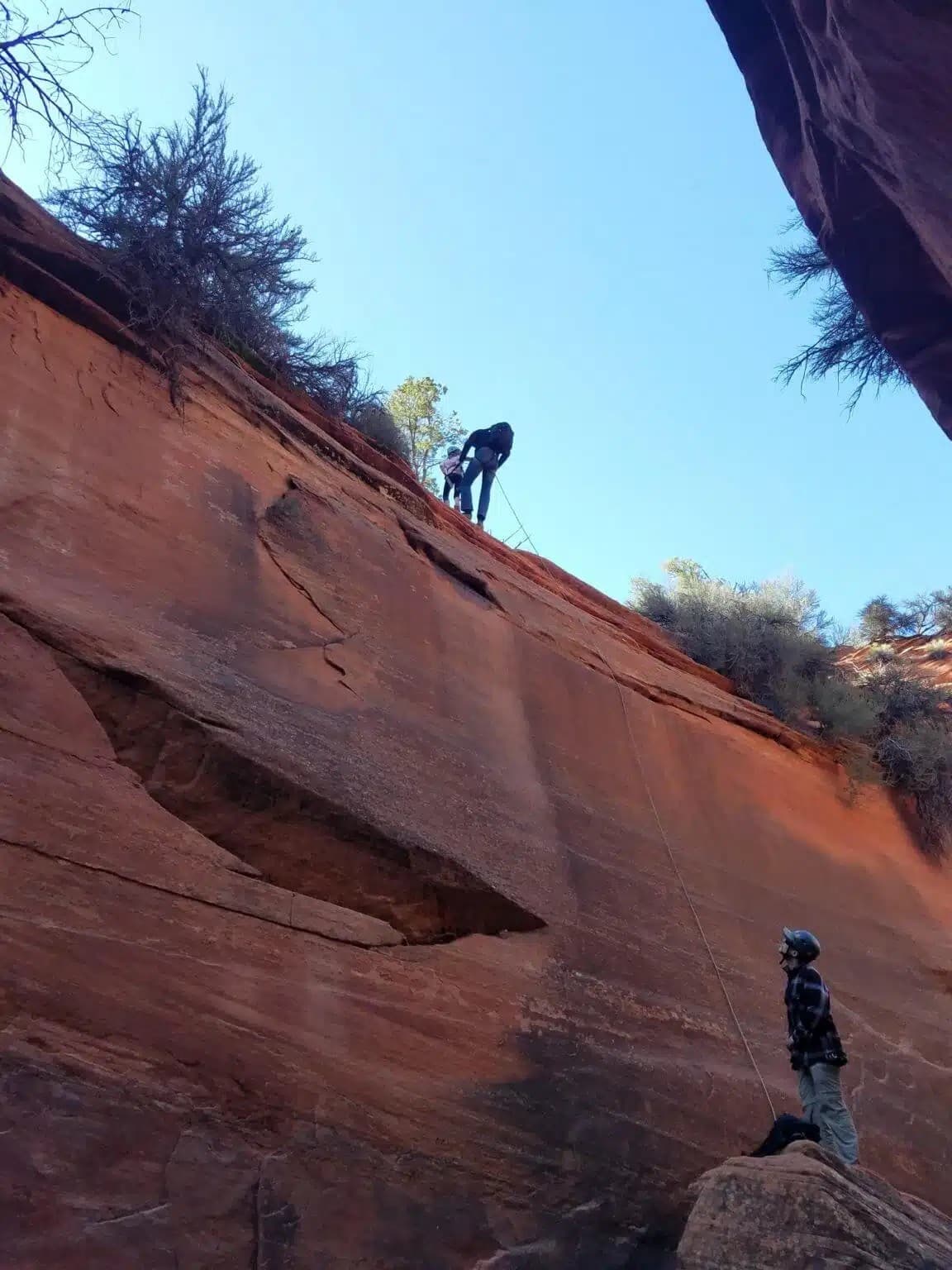 A person rappels down a steep red rock face while another individual watches from below.