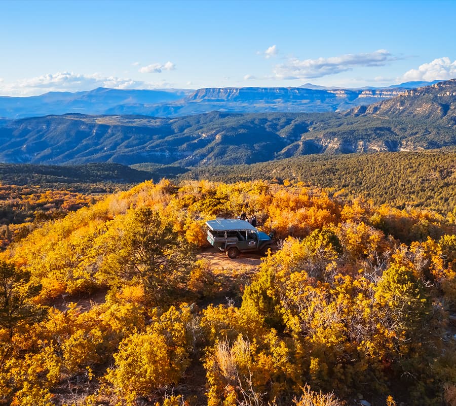 A black vehicle parked on a hillside surrounded by vibrant autumn foliage with a mountainous backdrop.