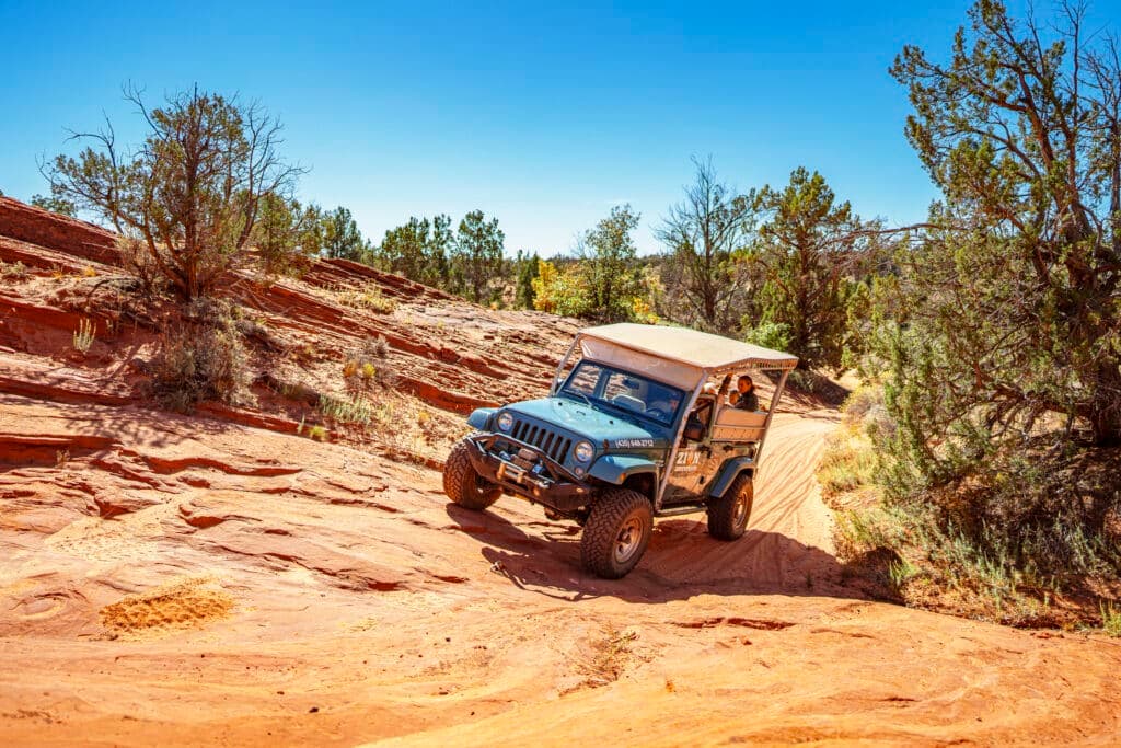 A blue Jeep navigates a rocky, sandy terrain under a clear blue sky.