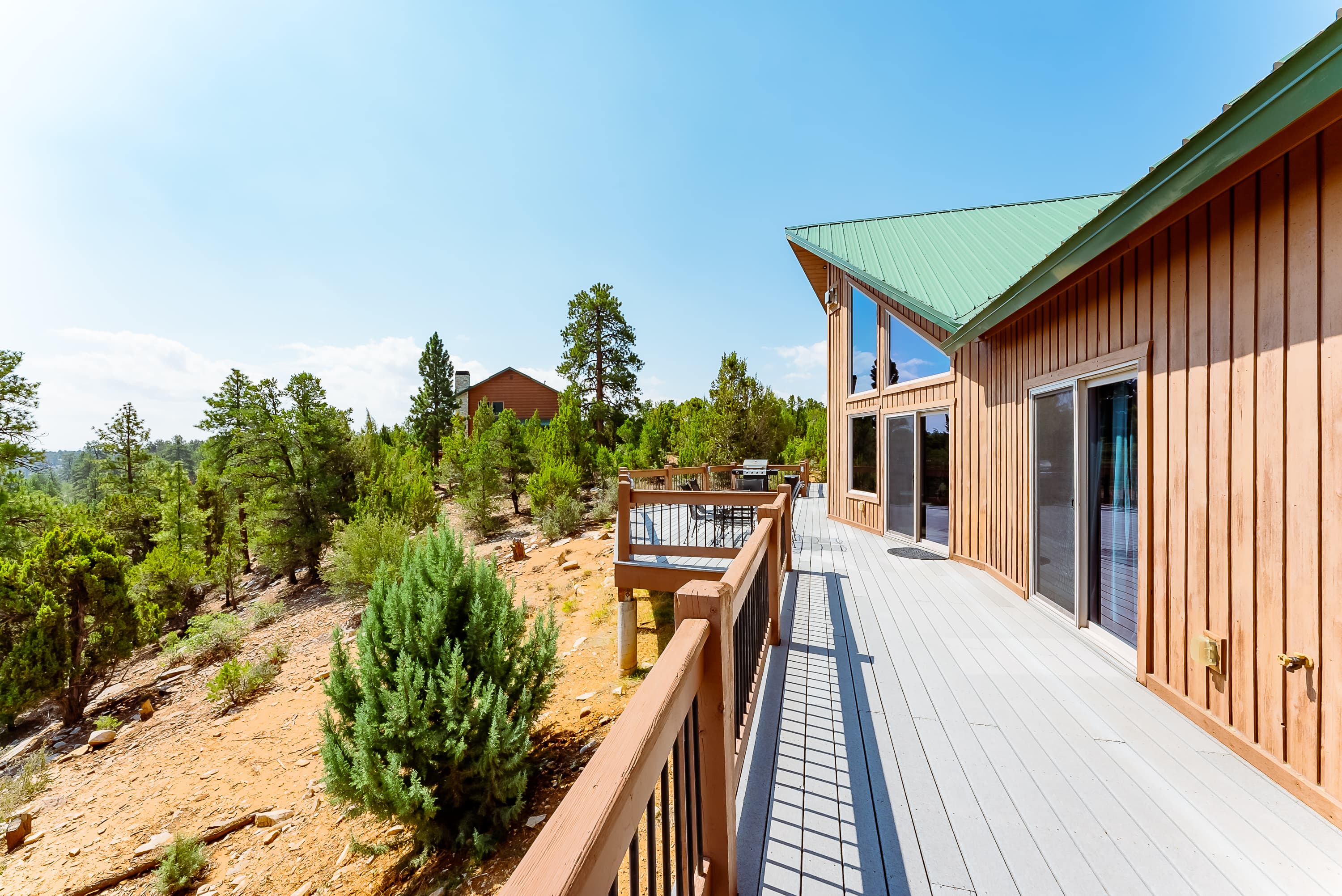 A wooden deck extends from a modern cabin, surrounded by pine trees and blue skies.