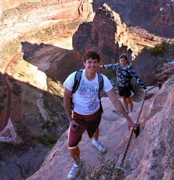 Two hikers navigate a steep, rocky trail with a chain for support against a backdrop of canyon cliffs.