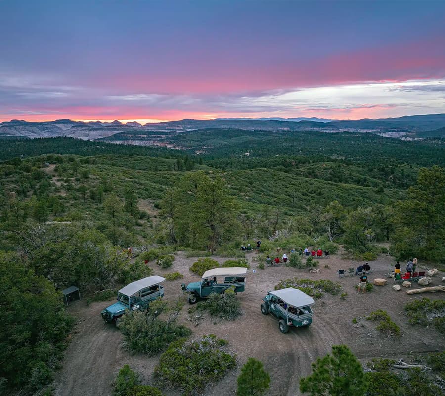 Aerial view of off-road vehicles parked on a hillside with a sunset and gathered group of people in the background.