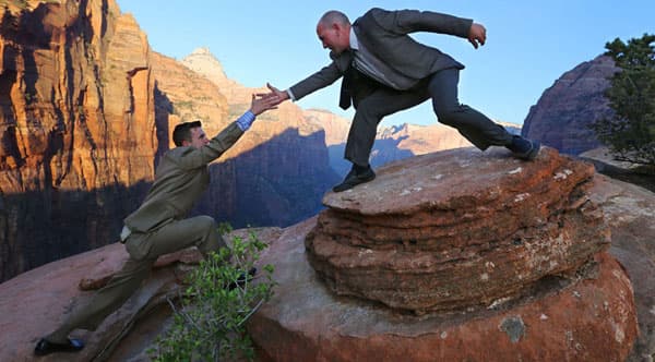 Two men in business suits reaching out to each other on a rocky cliff.