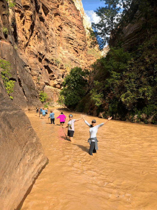 Hikers wade through a shallow river flanked by towering rock formations in a scenic canyon.