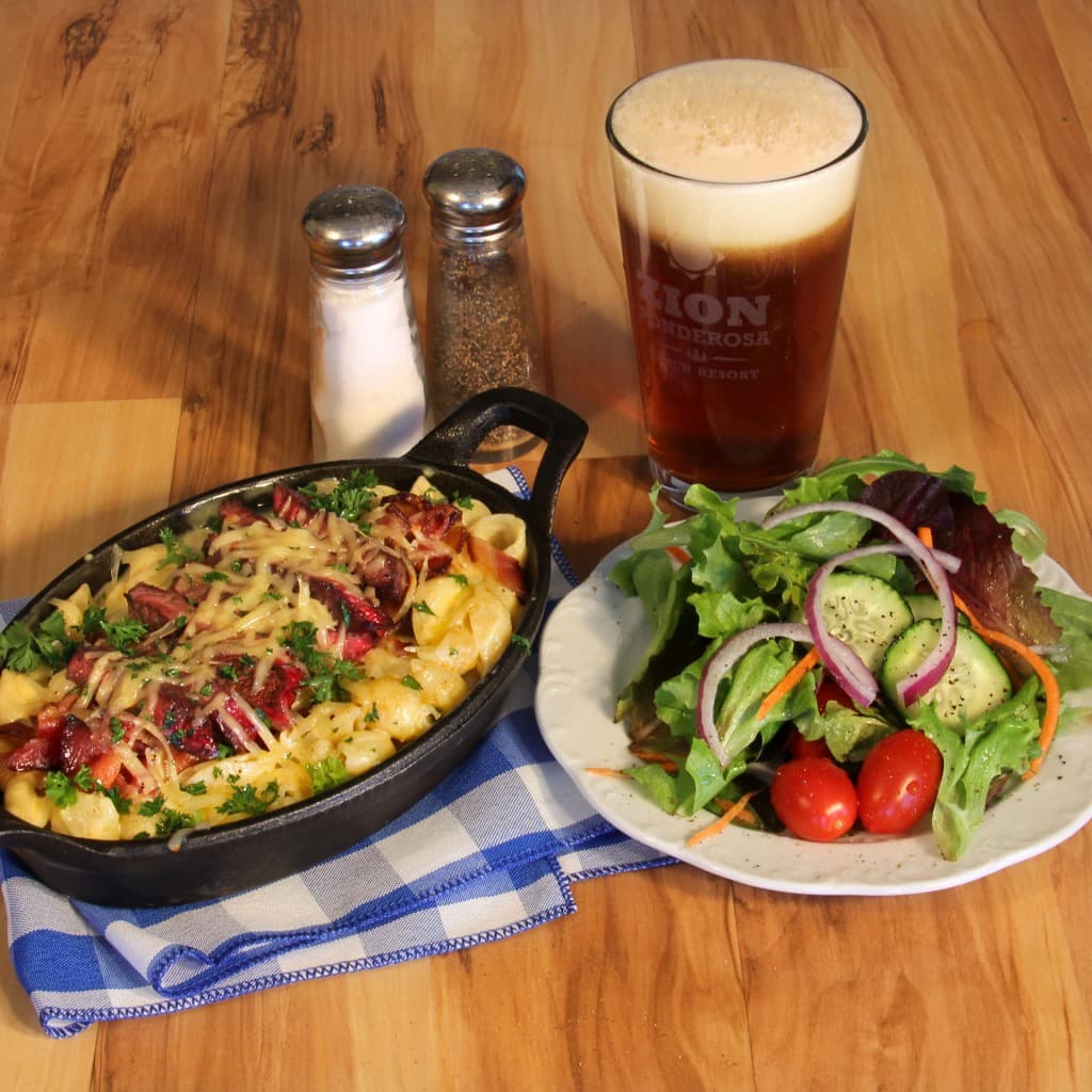 A serving of cheesy pasta with bacon and herbs, accompanied by a fresh salad and a glass of beer, all on a wooden table.