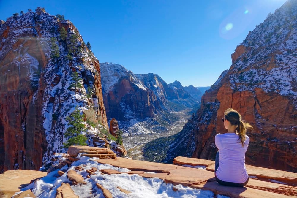 A person sits on a rocky ledge, gazing at a snowy canyon landscape under a clear blue sky.