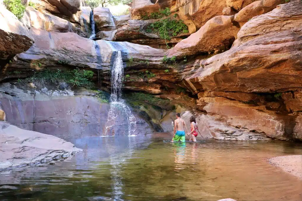 A couple stands in a serene pool of water beneath a waterfall surrounded by rocky cliffs and greenery.