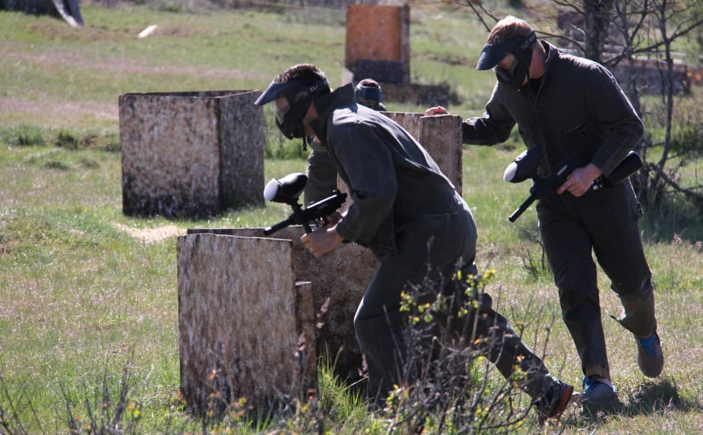 Two people in protective gear engage in a paintball match behind wooden obstacles in an outdoor setting.
