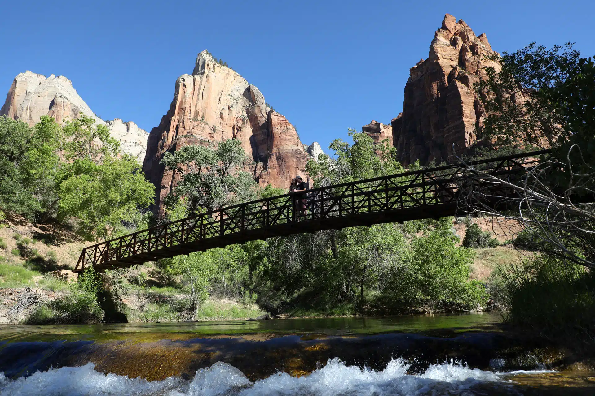 A wooden bridge spans a river with towering red rock formations in the background under a clear blue sky.