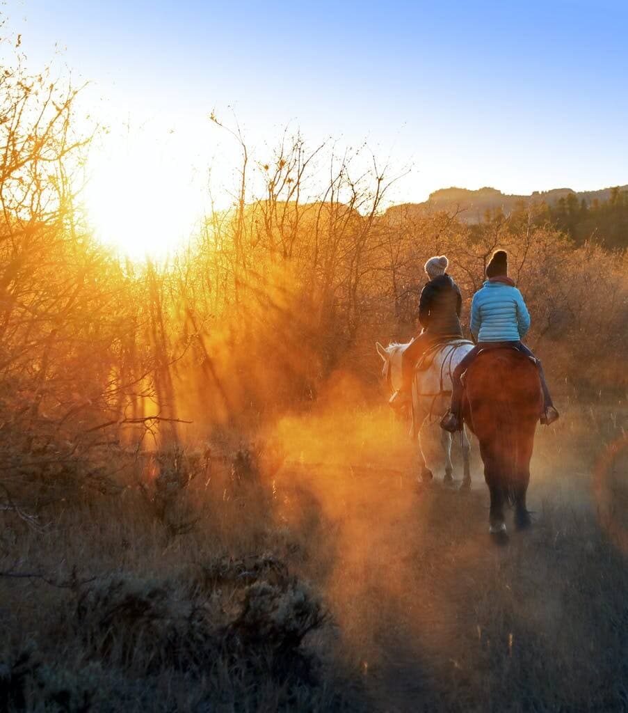 Two people riding horses along a misty path at sunrise.