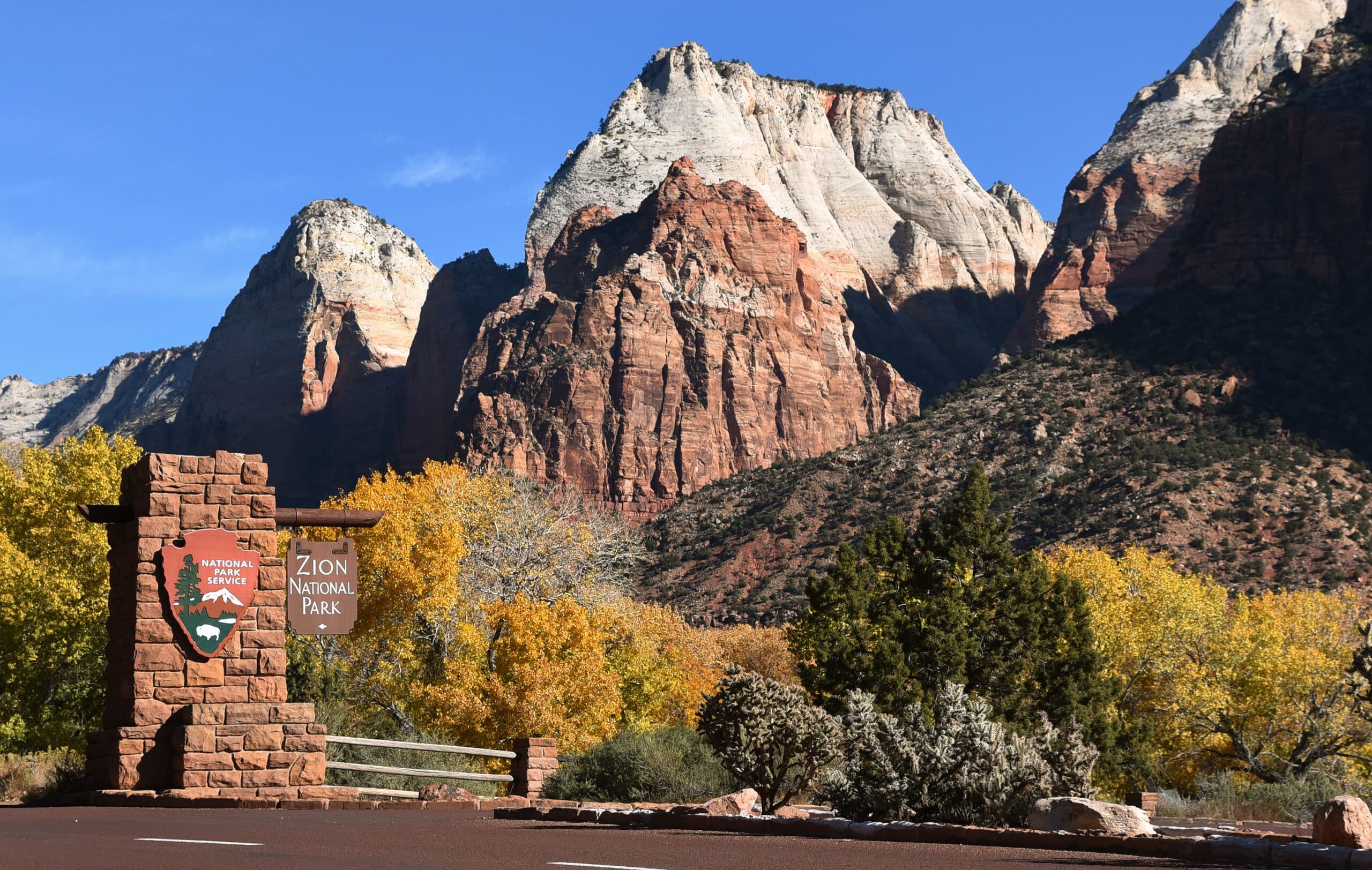 Entrance sign of Zion National Park with colorful autumn foliage and towering rock formations in the background.
