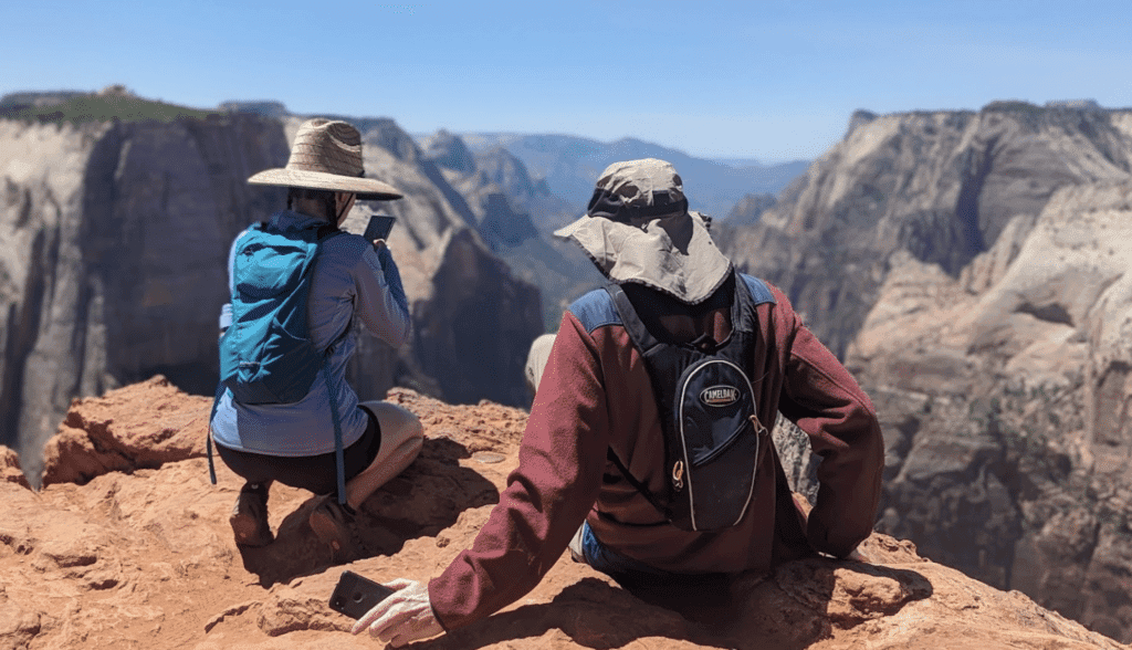 Two hikers sit on a rocky ledge, one photographing the canyon view while the other relaxes.
