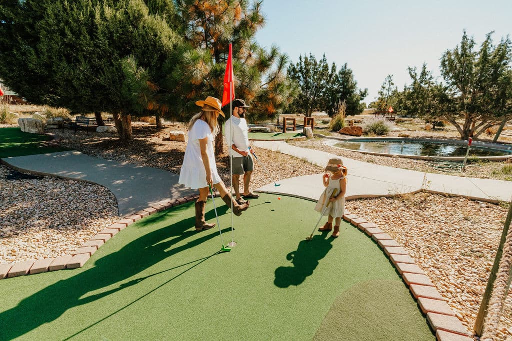 A family enjoys a game of mini-golf on a sunny day, with a child watching nearby.