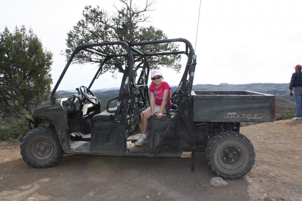 A girl in a pink shirt sits on the side of an all-terrain vehicle parked on a rocky terrain with mountains in the background.