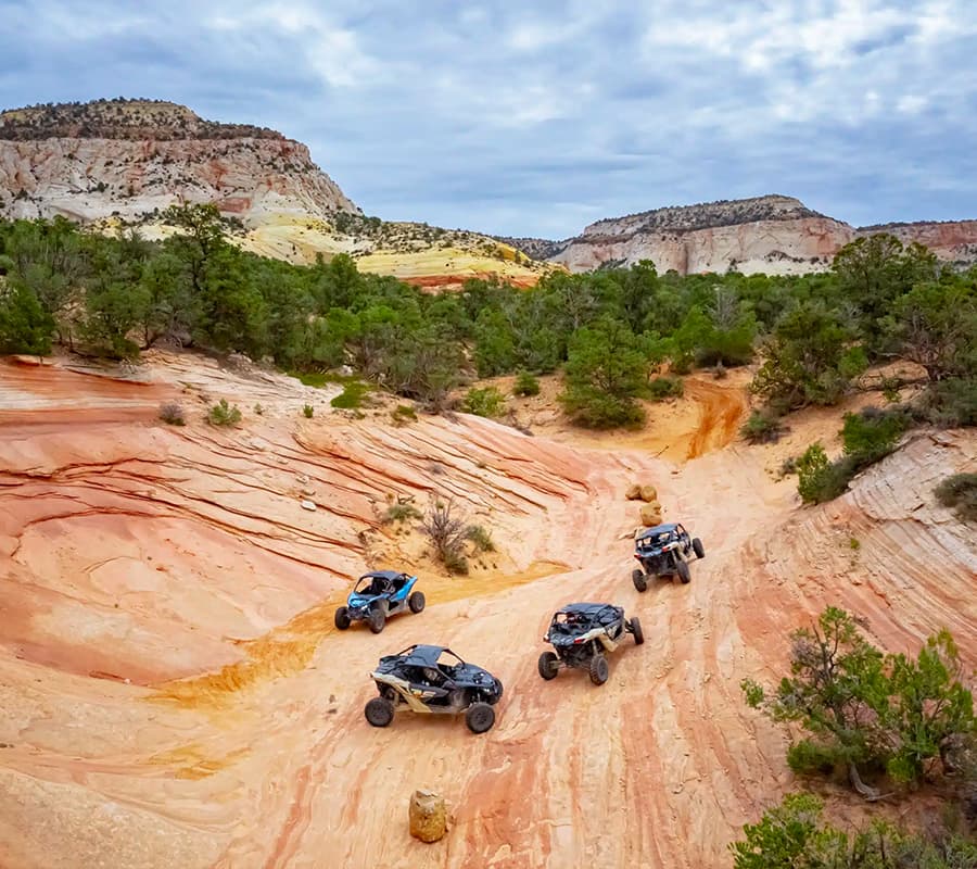 Four off-road vehicles navigate a winding, sandy path through colorful rock formations and greenery.