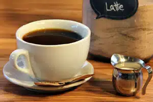A white cup filled with coffee beside a small metal creamer and a bag labeled "Latte."