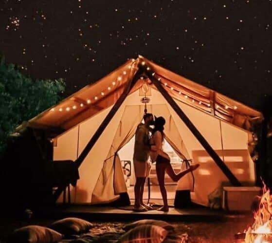 A couple kisses in front of a lit tent under a starry night sky.