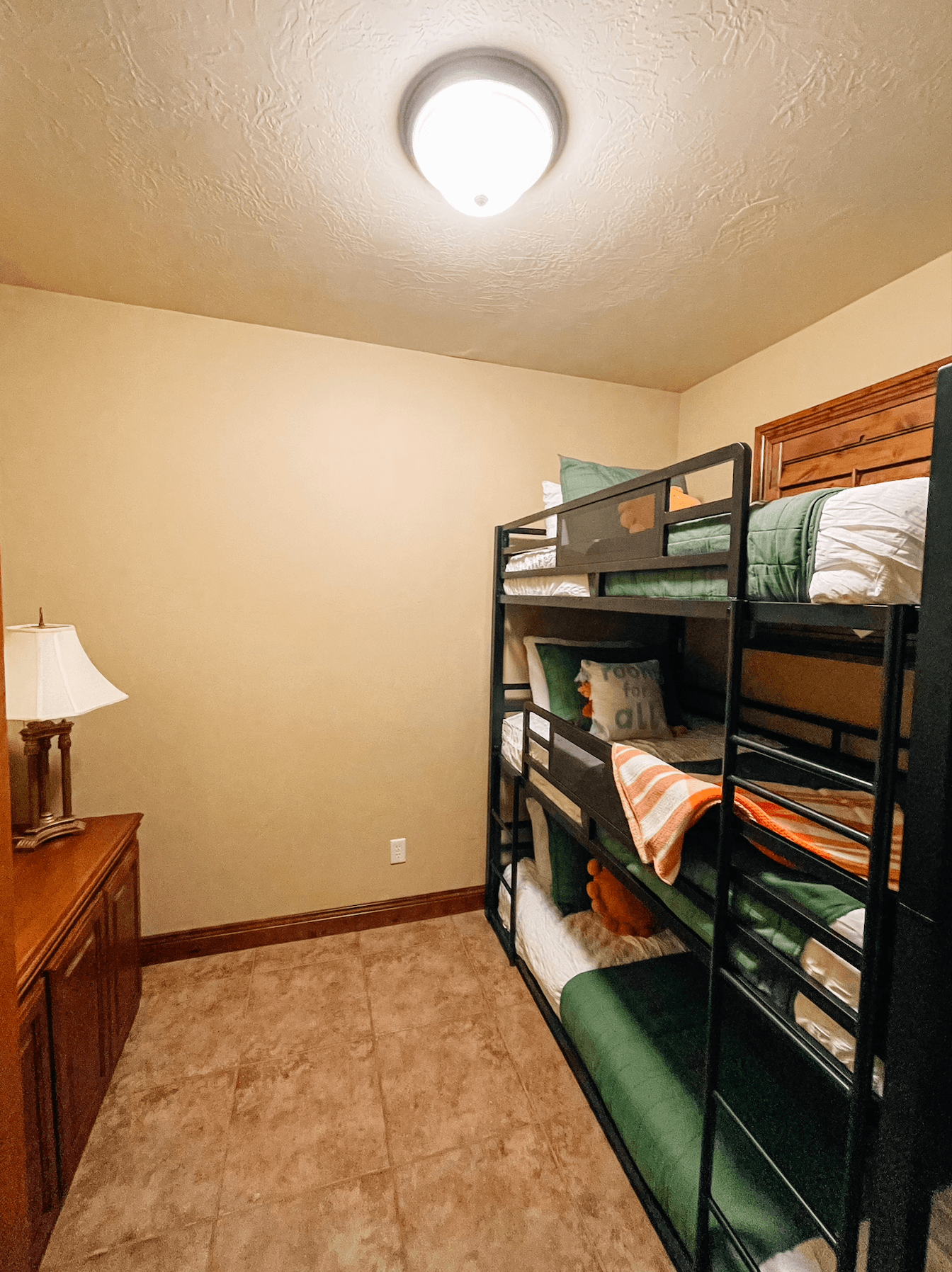 A small room featuring a black metal bunk bed with green bedding, a lamp, and a wooden dresser.