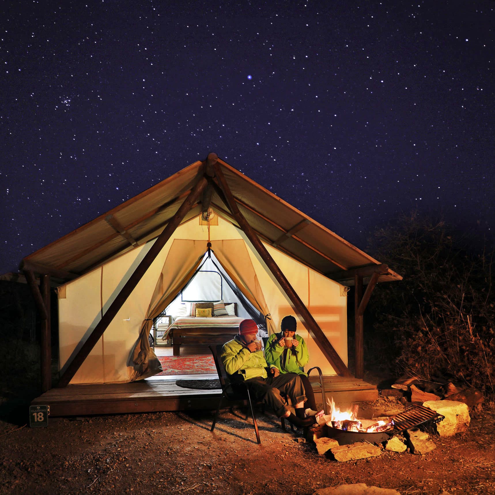 Two people sit by a campfire in front of a glamping tent under a starry night sky.