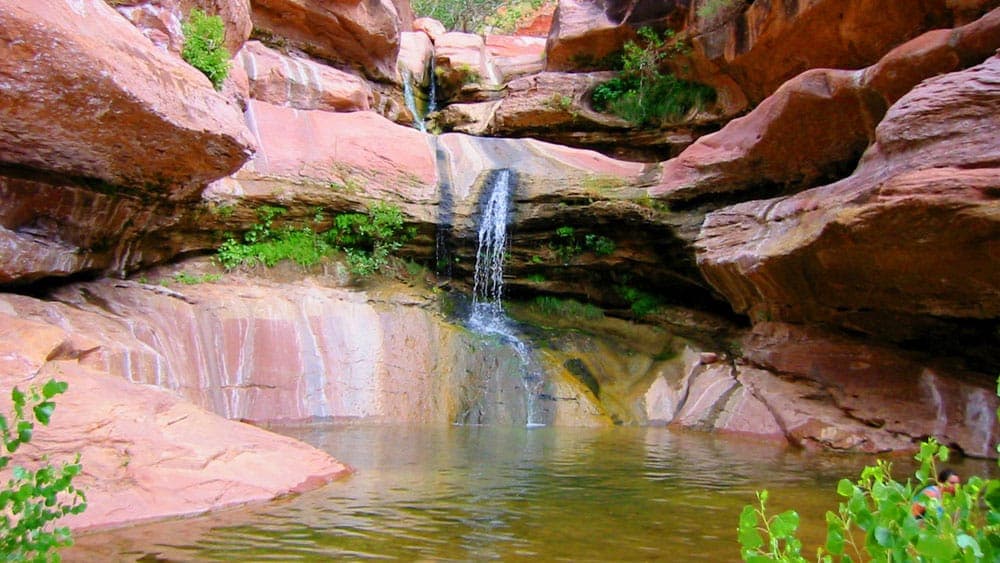 A serene waterfall flowing over rock formations into a clear pool surrounded by greenery.