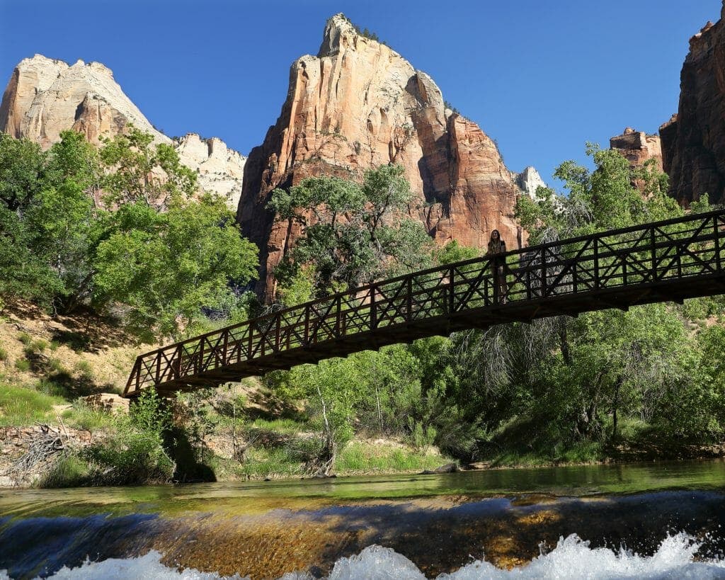 A wooden bridge spans a river surrounded by green foliage and towering rock formations under a clear blue sky.