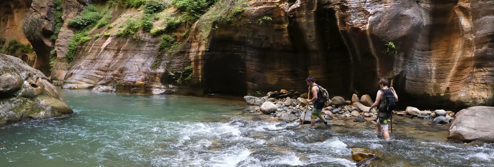 Two hikers wade through a rocky river surrounded by tall, rugged canyon walls.