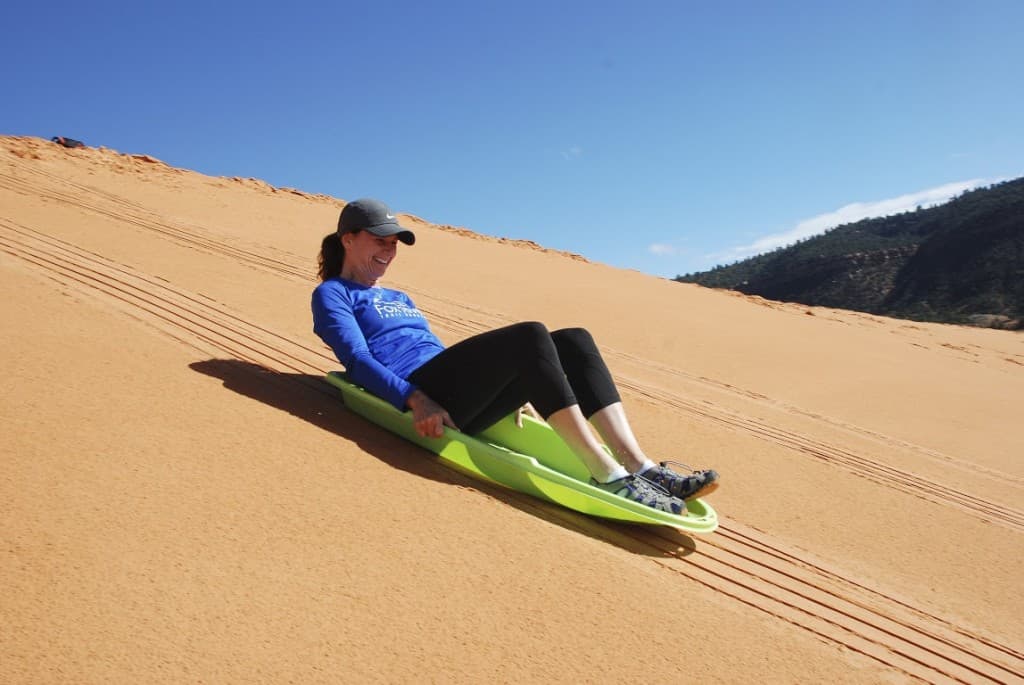 A person in a blue shirt slides down a sandy dune on a green sled.