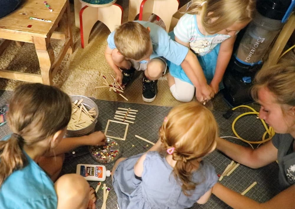Children engaged in a crafting activity with popsicle sticks and beads on a carpeted floor.
