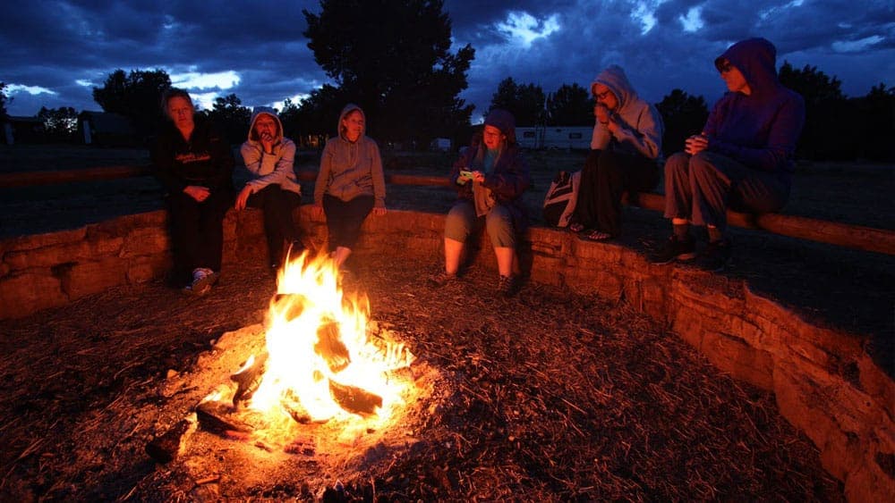 A group of people gathers around a warm campfire under a darkening sky.